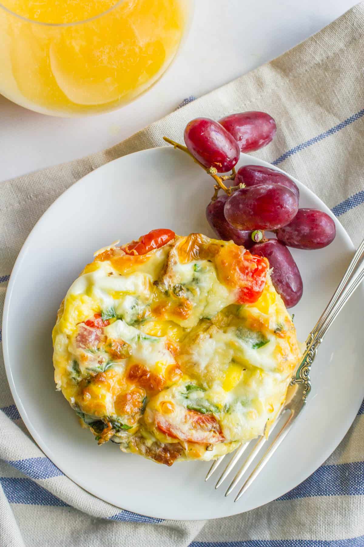 A fork resting next to an individual egg and vegetable breakfast casserole with some red grapes to the side.