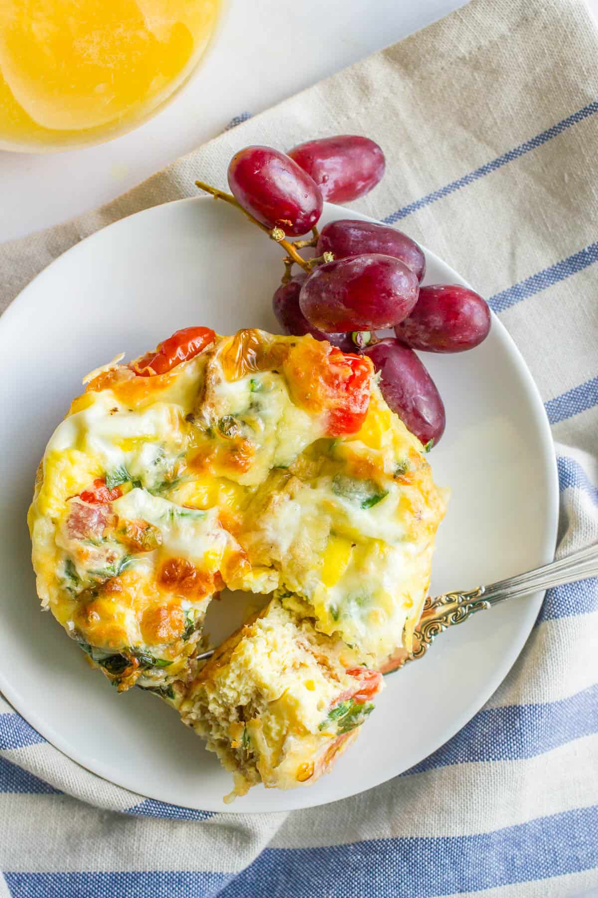 A fork holding a piece of a breakfast casserole on a white round plate with red grapes.