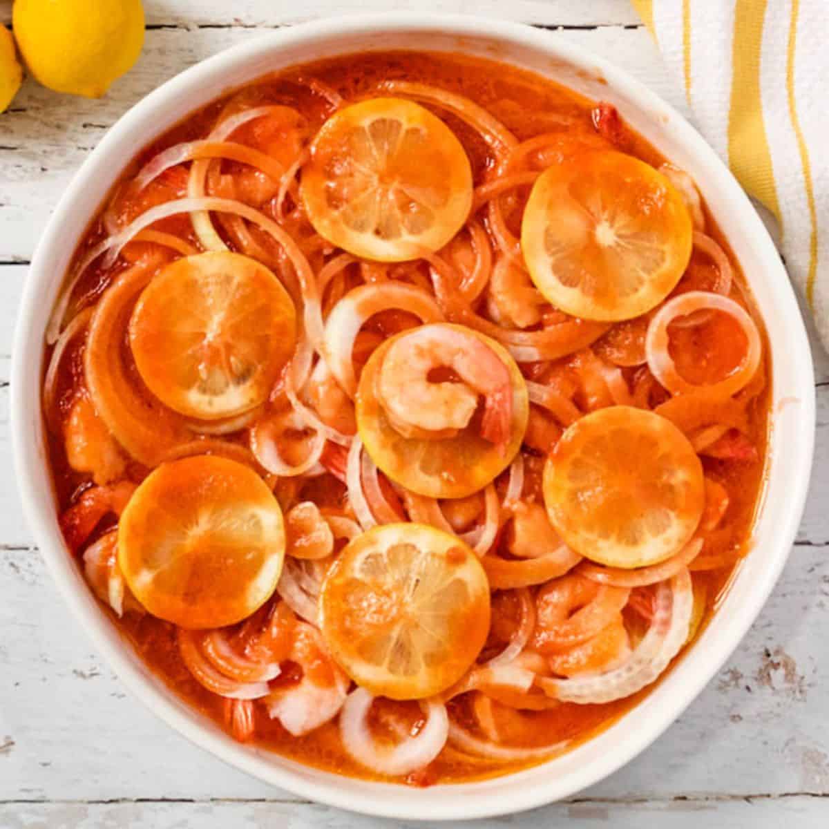 Marinated shrimp with sliced onions and lemons in a large white round casserole dish.