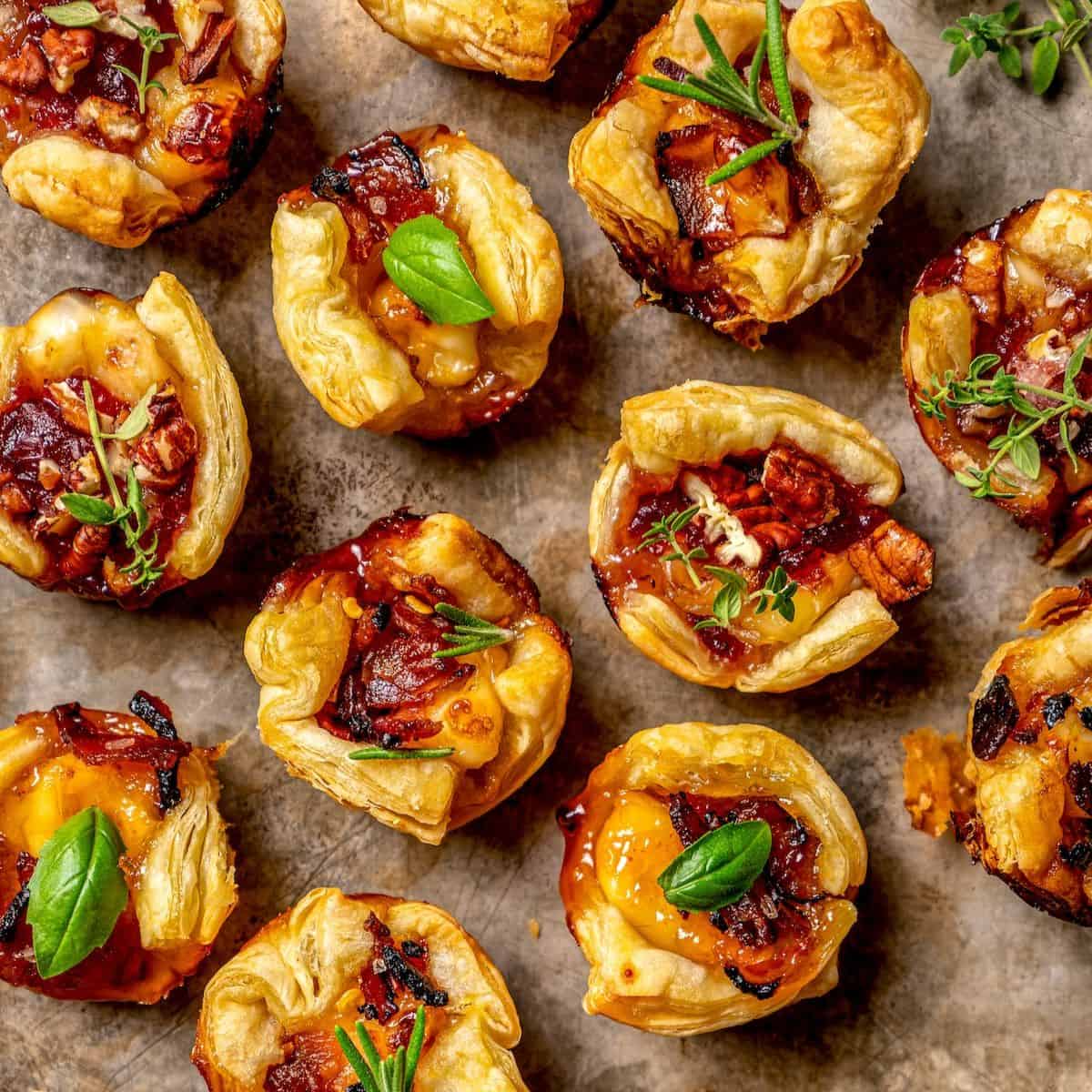 Close up of puff pastry brie bites with pecans, bacon and other toppings and sprigs of fresh herbs for garnish.