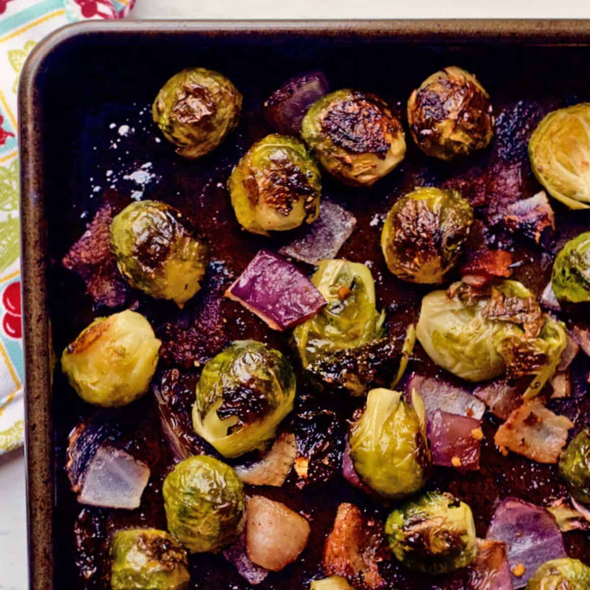 Roasted Brussels sprouts with bacon and red onion pieces on a dark sheet pan.