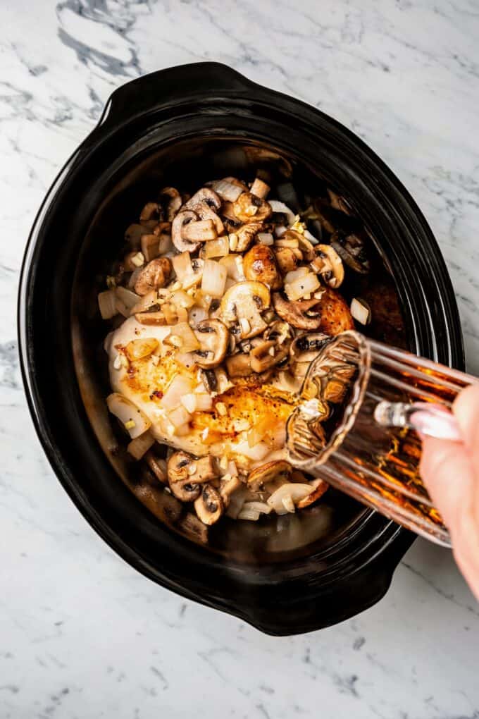 A marsala wine sauce being poured over chicken breasts in a crock pot insert.