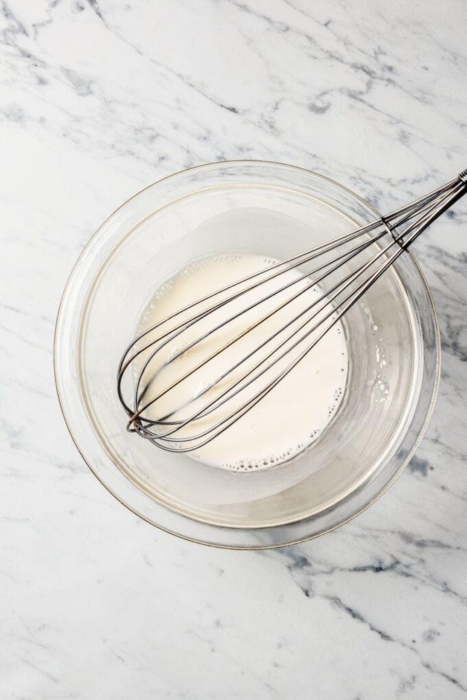 A whisk resting in a small glass bowl with a cornstarch slurry mixture in it.