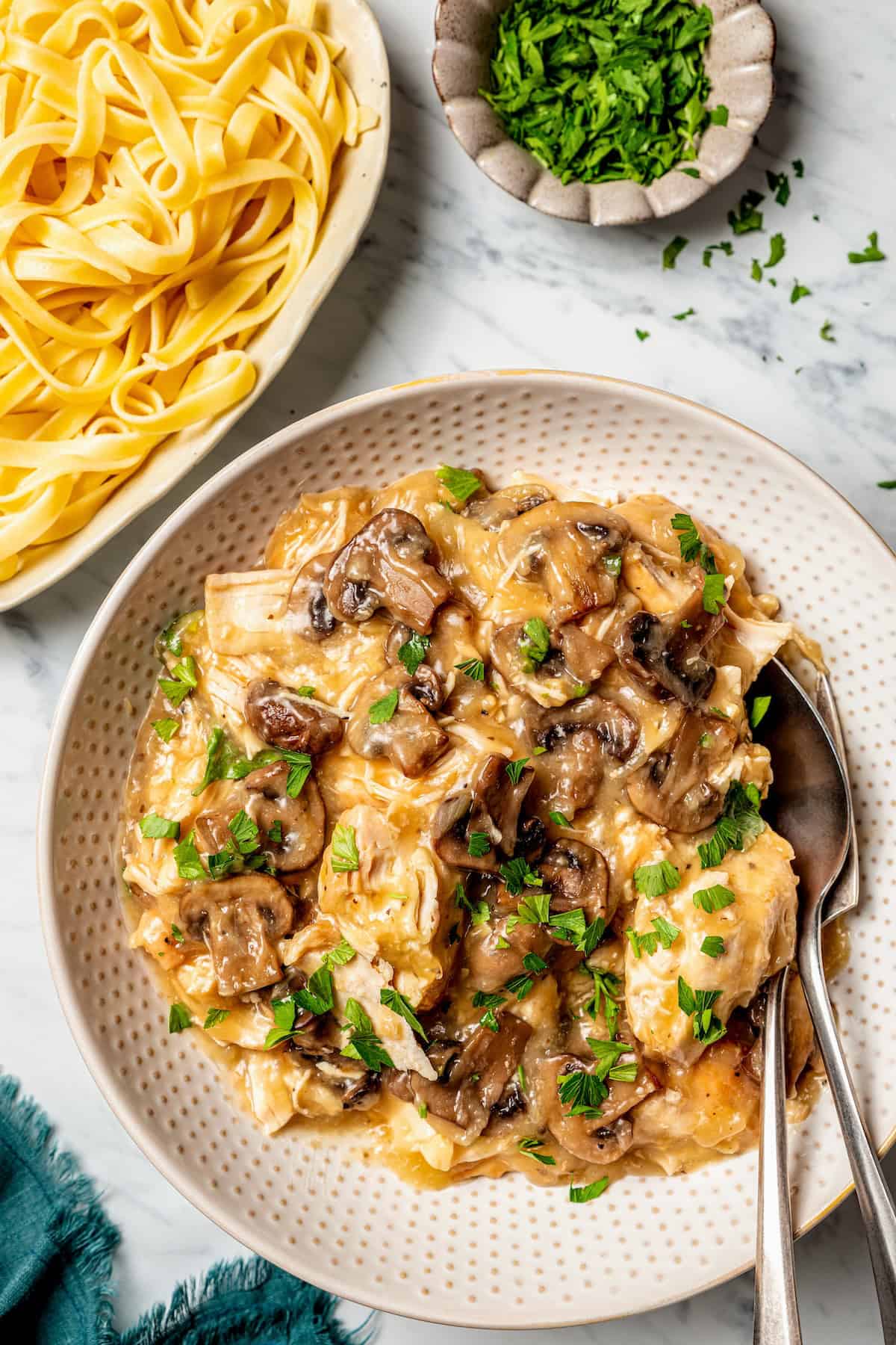Chicken marsala served in a large serving bowl with pasta to the side and serving utensils resting in the bowl.