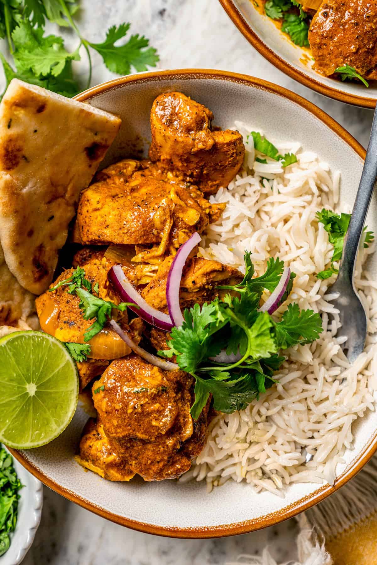 A fork resting in a bowl of rice and chicken tikka masala with naan bread to the side and a lime wedge for garnish.