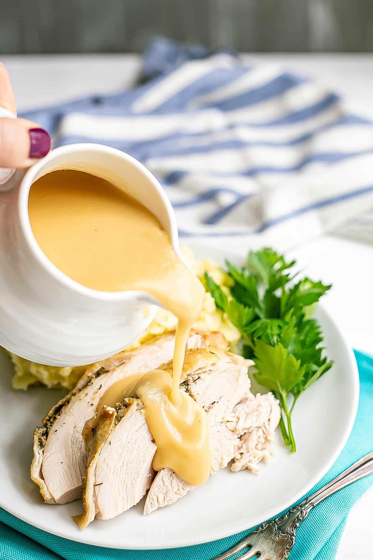 Gravy being drizzled over slices of slow cooker turkey breast served with mashed potatoes on a white plate.