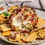 A cheese ball with bacon, cheese, green onions, nuts and dried cranberries on a plate with crackers and a knife resting to the side and a text overlay on the photo.