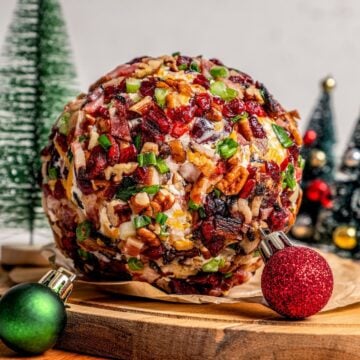Close up of a cheese ball appetizer on a cutting board with Christmas decorations in the background.