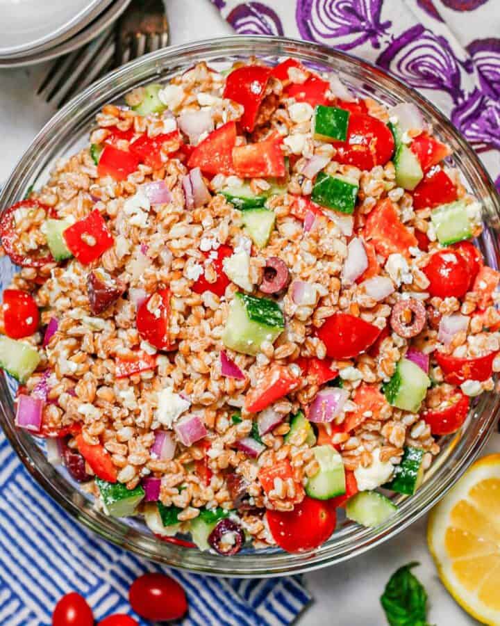 Close up of Mediterranean farro salad with olives and feta cheese in a large glass bowl with colorful throw towels nearby.