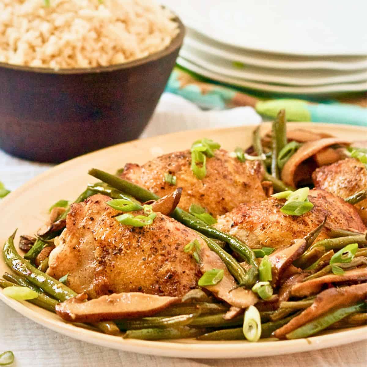 Close up of a platter of seared chicken thighs with shiitake mushrooms and French green beans with a bowl of rice in the background.