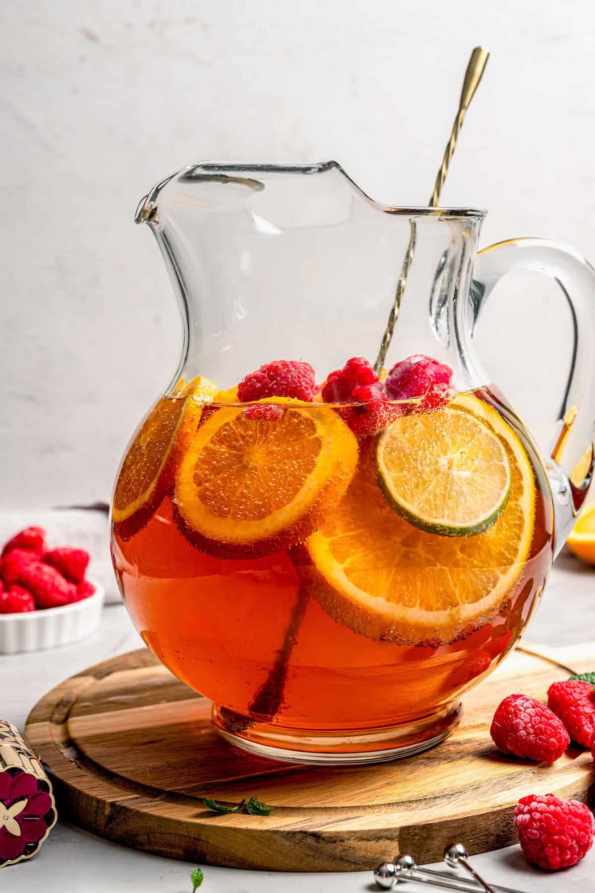 A glass pitcher full of a bubbly holiday punch with slices of orange and lime and fresh raspberries.