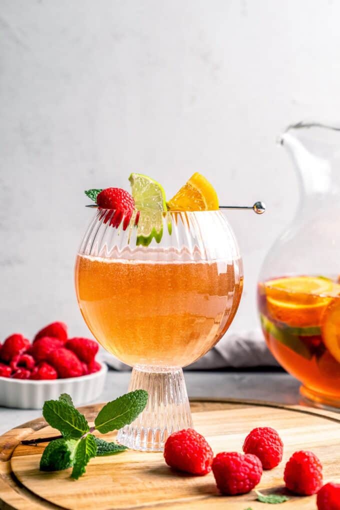 A glass with a bubbly punch mixture topped with fresh fruit and a scattering of raspberries to the side.