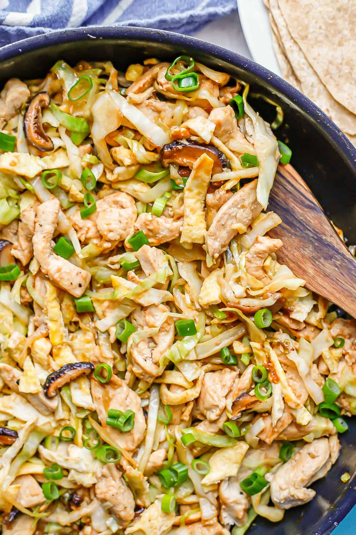 A wooden spatula resting in a skillet of moo shu chicken with cabbage and mushrooms.