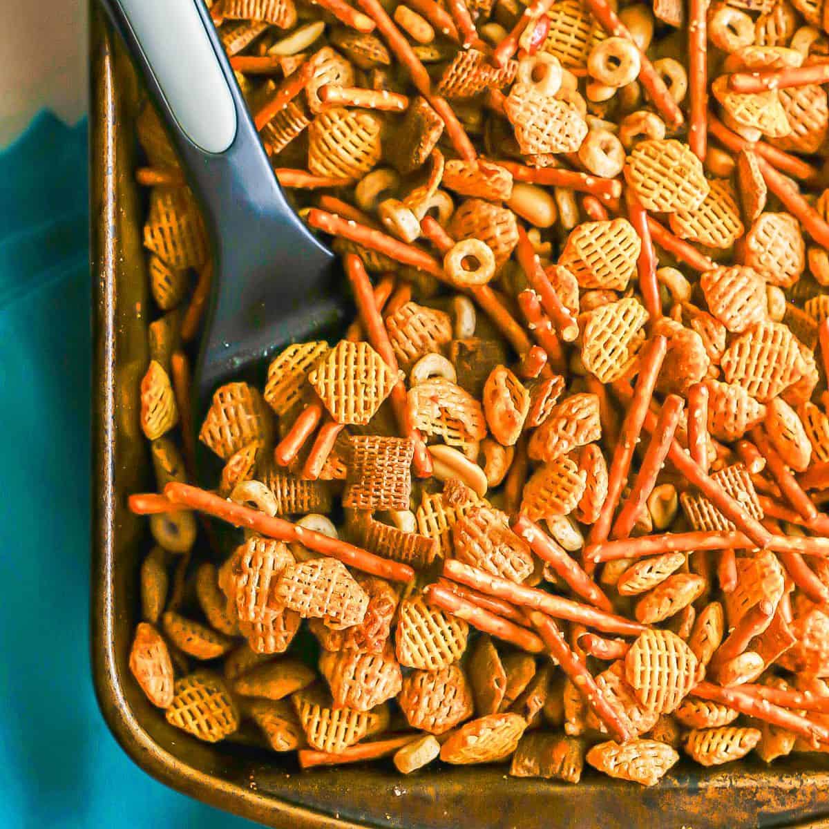 Close up of a spatula resting in a rectangular baking pan full of a chex mix mixture.