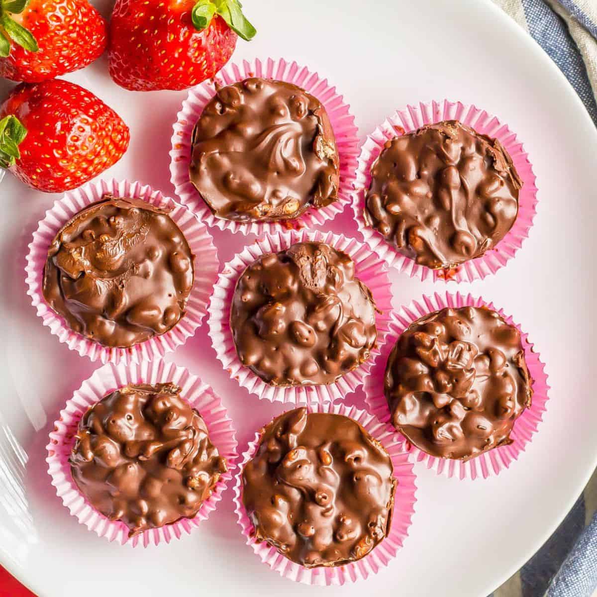 Seven mini homemade candies in pink cupcake liners on a white plate with strawberries to the side.
