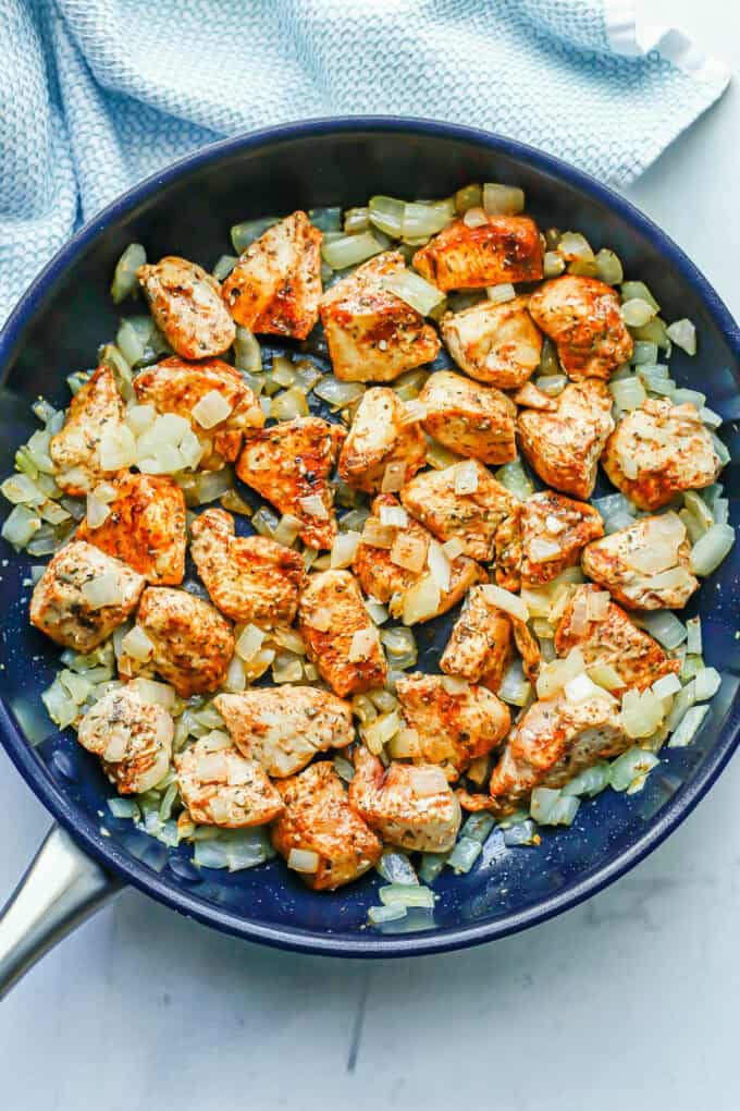 Chunks of chicken seasoned and cooking in a large skillet with sautéed onions.