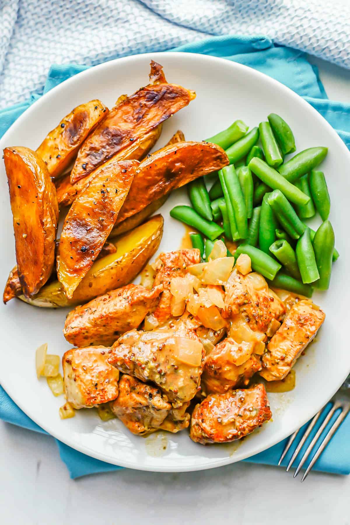 Cubed chicken with onions and a honey mustard sauce served on a dinner plate with green beans and potato wedges.