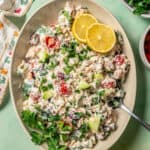 An oval serving platter with a veggie loaded Mediterranean tuna salad garnished with fresh herbs and lemon slices and a serving spoon resting in it and a text overlay on the photo.