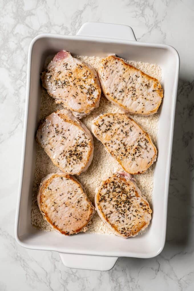 Seared pork chops atop white rice in a casserole dish before being cooked.