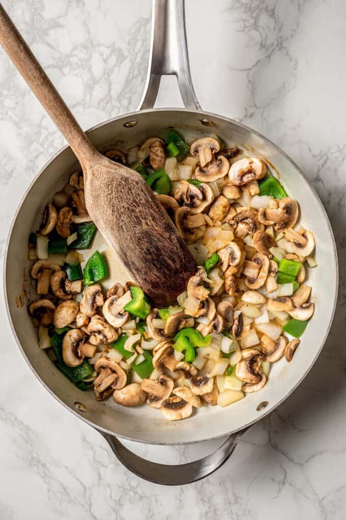 A skillet with a mixture of onions, green peppers and mushrooms and a wooden spoon resting in it.