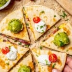 A hand picking up a slice of a bacon egg and cheese breakfast quesadilla cut into wedges and served with guacamole and sour cream with a text overlay on the photo.