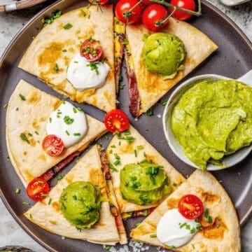 Close up of wedges of breakfast quesadillas served on a dark plate with guacamole and sour cream on top and chipped fresh chives sprinkled around.
