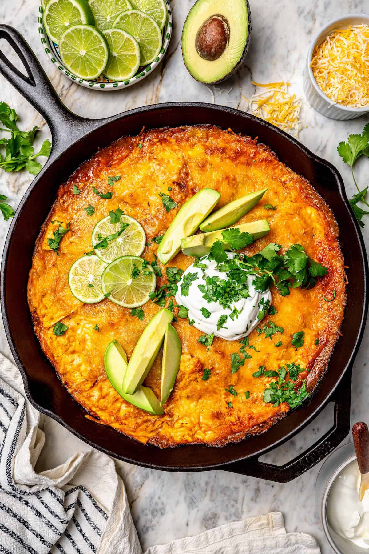 Chicken enchilada skillet in a cast iron pan with lime wedges, avocado slices, sour cream and chopped fresh herbs on top.
