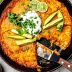 A spatula resting in a cast iron pan with a chicken enchilada mixture topped with lime wedges, avocado slices, sour cream and chopped fresh herbs with a text overlay on the photo.