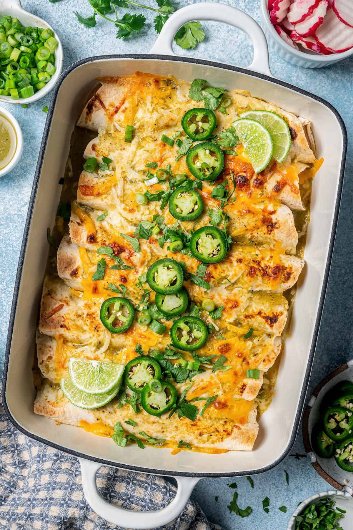 A casserole dish with baked cream cheese chicken enchiladas topped with slices of jalapeños, lime and chopped fresh green onions.