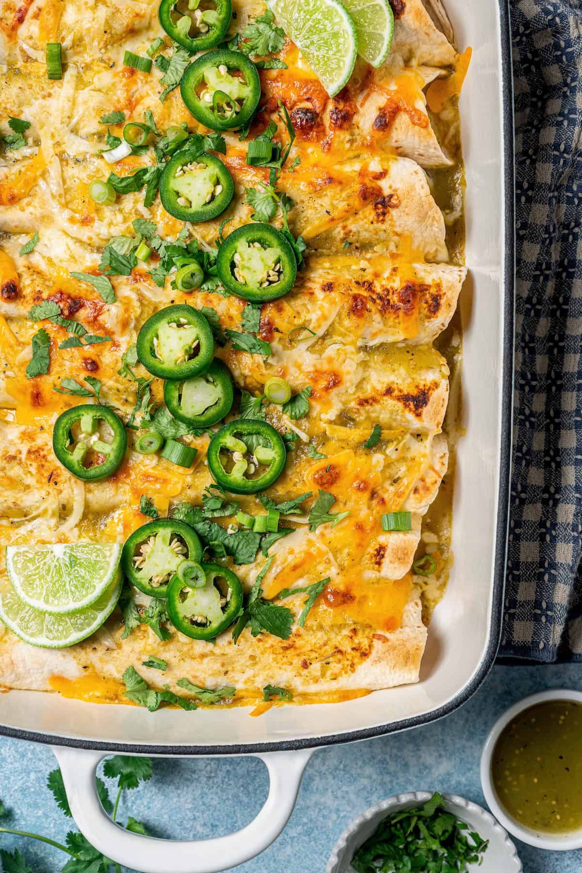 Close up of a casserole dish with baked cream cheese chicken enchiladas topped with slices of jalapeños, lime and chopped fresh green onions.