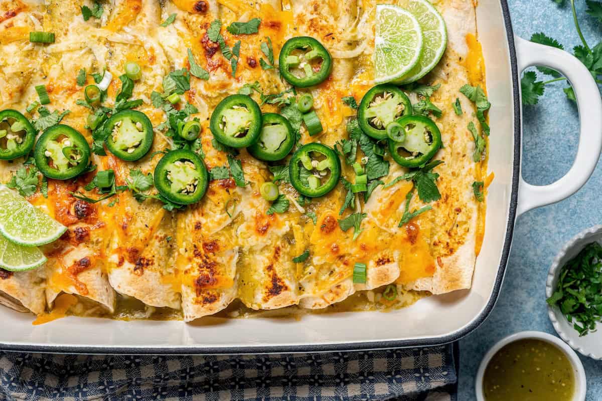 Close up of a casserole dish set on a blue and white napkin with baked cream cheese chicken enchiladas topped with slices of jalapeños, lime and chopped fresh green onions.