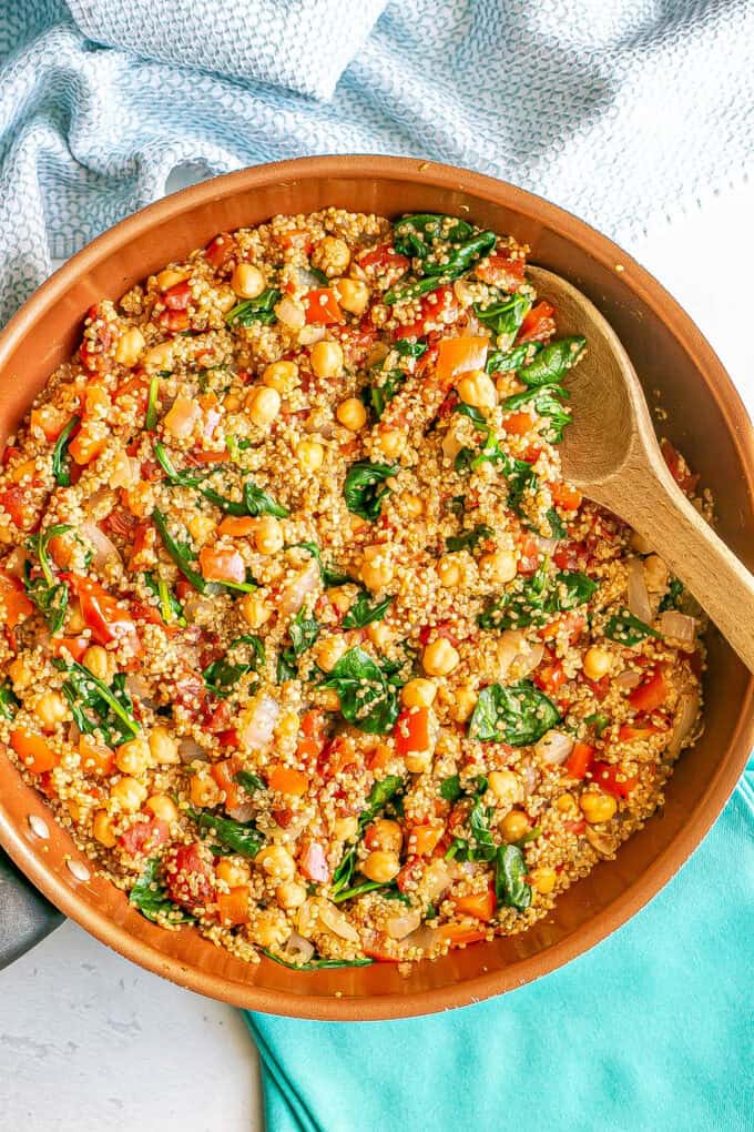 A copper skillet with a cooked quinoa, spinach, tomatoes and chickpea mixture and a wooden spoon resting in it.