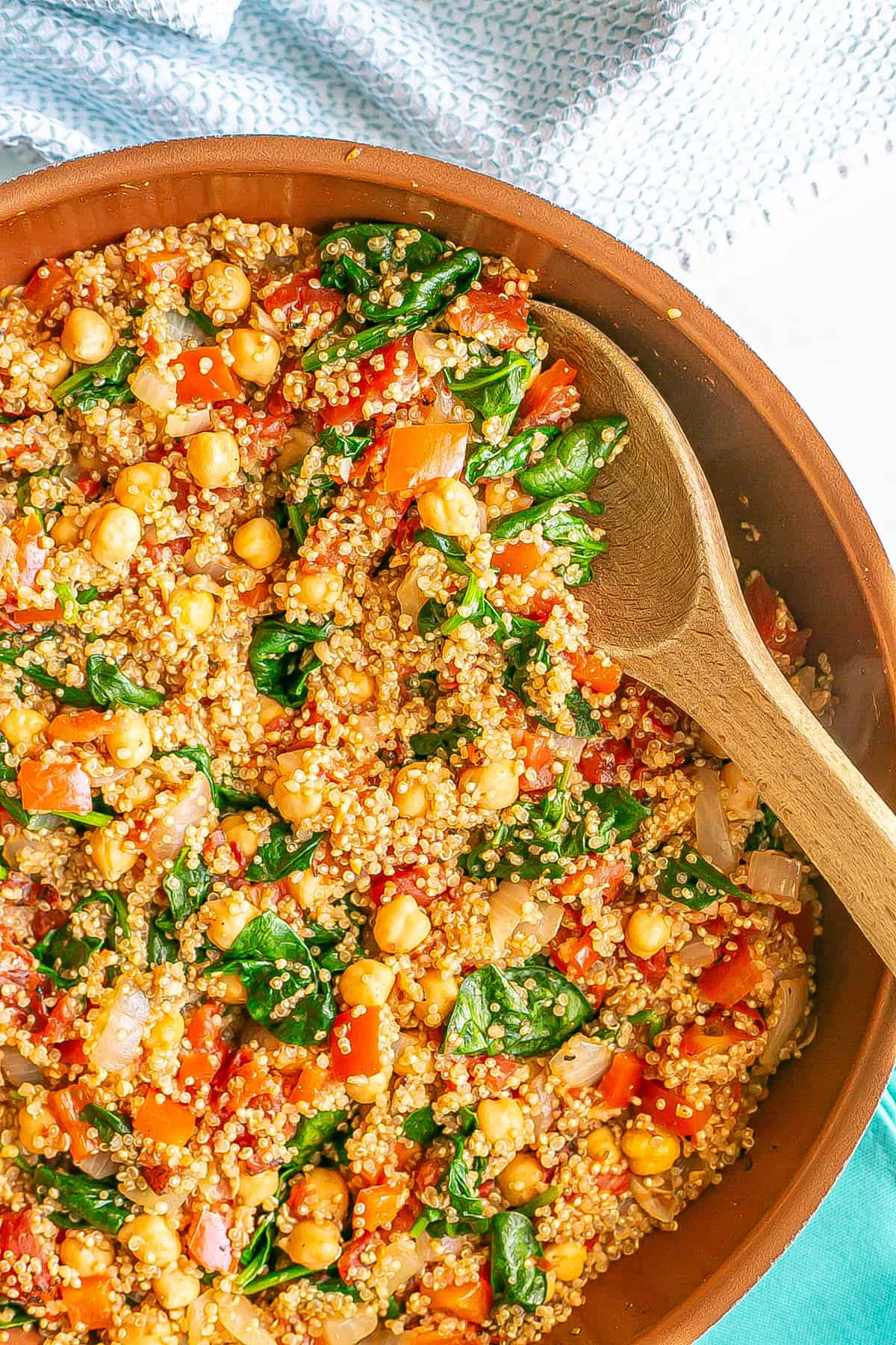 Close up of a wooden spoon resting in a copper skillet with a cooked quinoa, spinach, tomatoes and chickpea mixture.