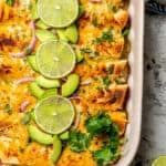 Close up of baked cheesy chicken enchiladas verde in a casserole dish with cilantro, lime and avocado scattered on top with a text overlay on the photo.