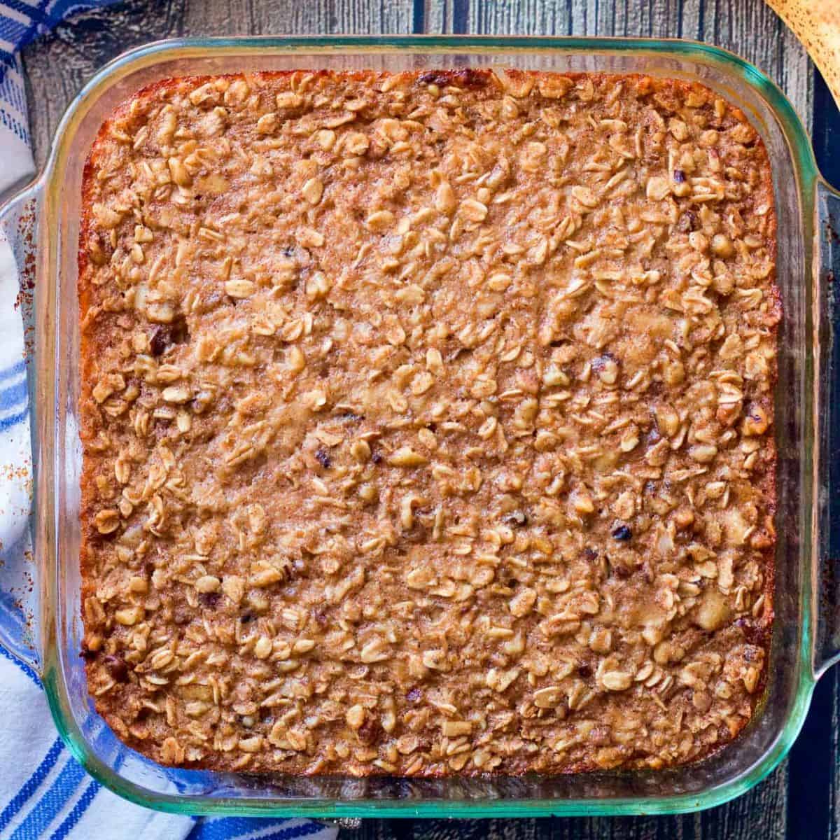 An 8x8 glass baking dish with a banana bread flavored baked oatmeal alongside a ripe banana and a blue striped kitchen towel.