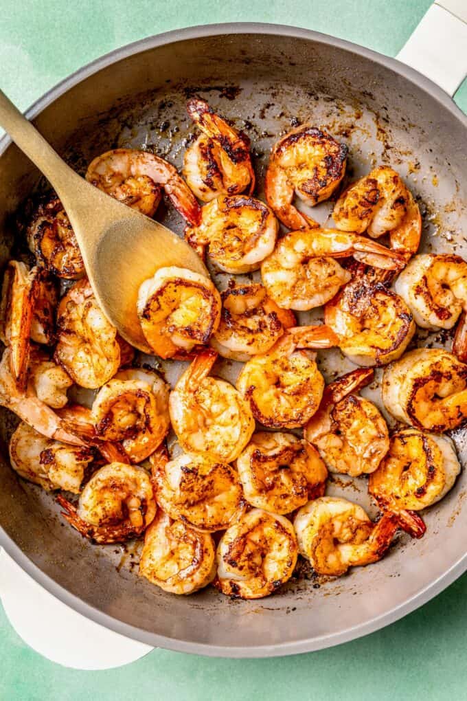 A wooden spoon resting in a skillet of cooked, charred shrimp.