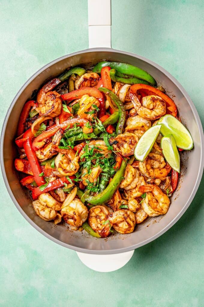 A skillet with cooked, charred shrimp, peppers and onion with cilantro sprinkled on top and some lime wedges on the side.