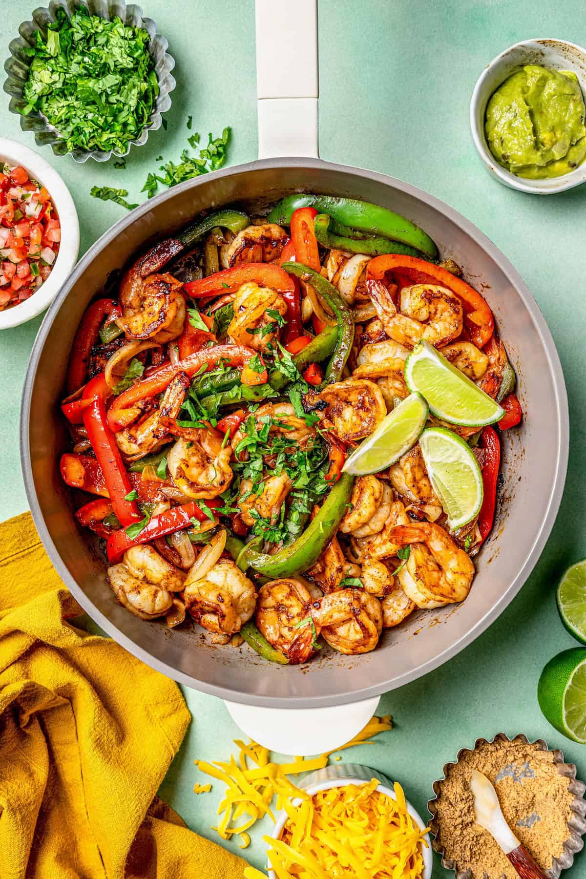 A skillet with cooked, charred shrimp, peppers and onion with cilantro sprinkled on top and lime to the side and surrounded by small bowls of toppings.