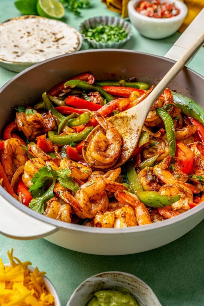 A spoon holding a cooked shrimp in a skillet of shrimp fajitas.