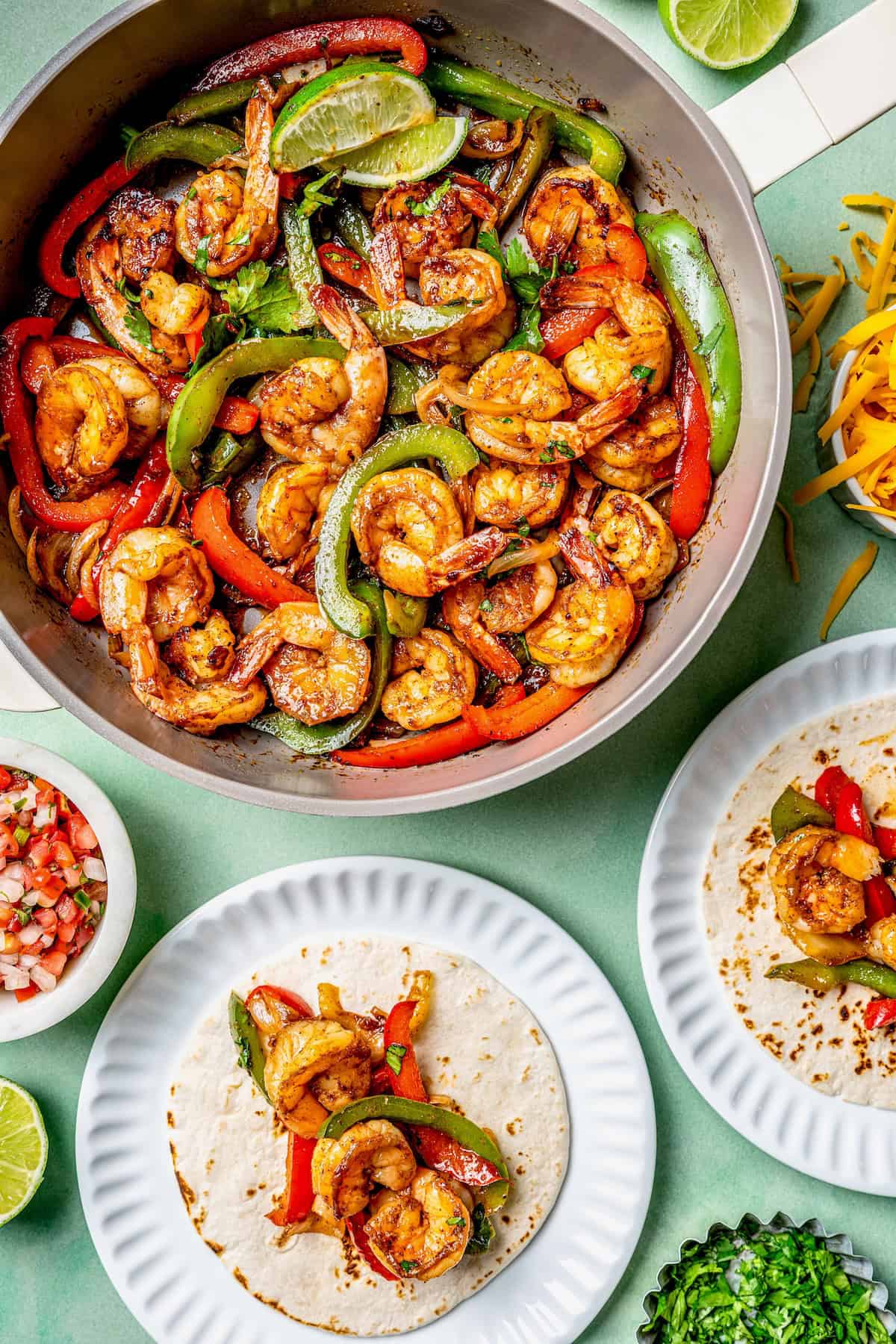 A skillet full of shrimp fajitas alongside plates with tortillas topped with the fajita mixture.