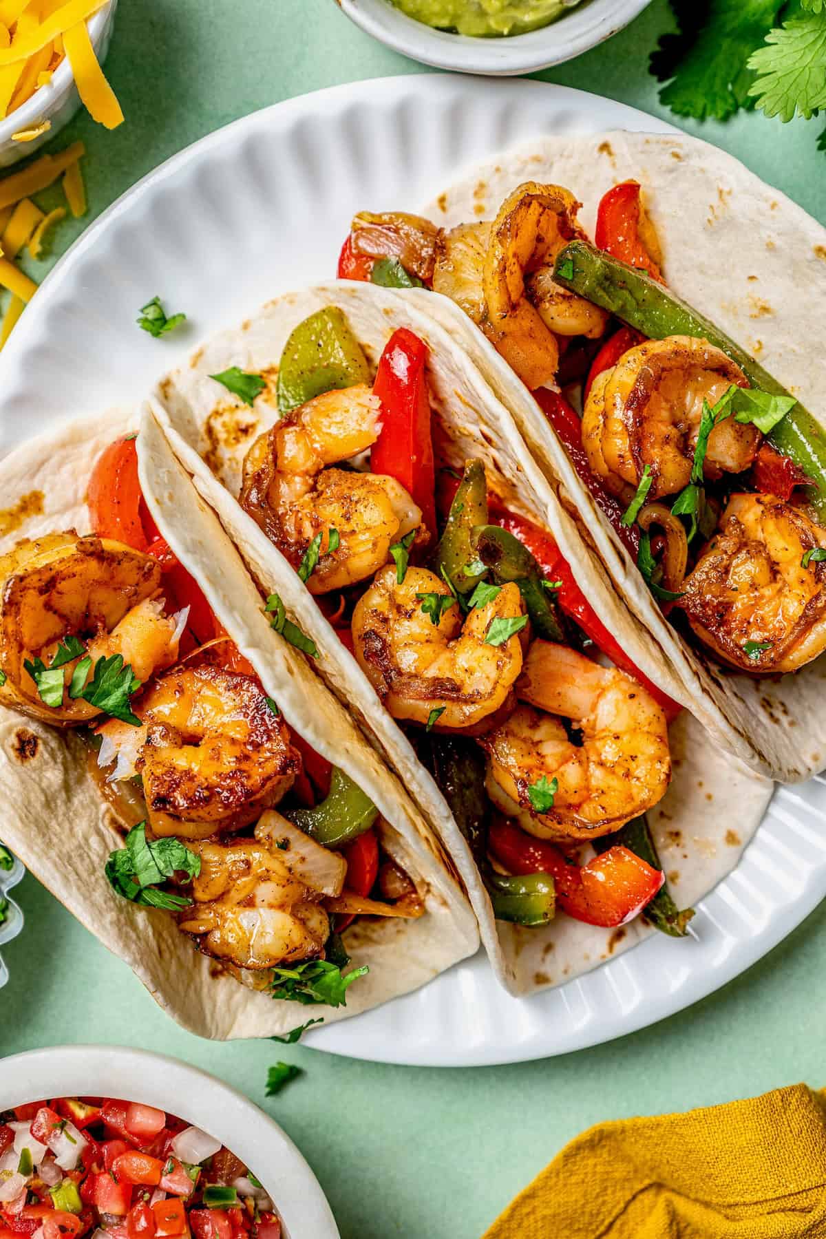 Three shrimp fajitas on a white plate with toppings in bowls nearby.