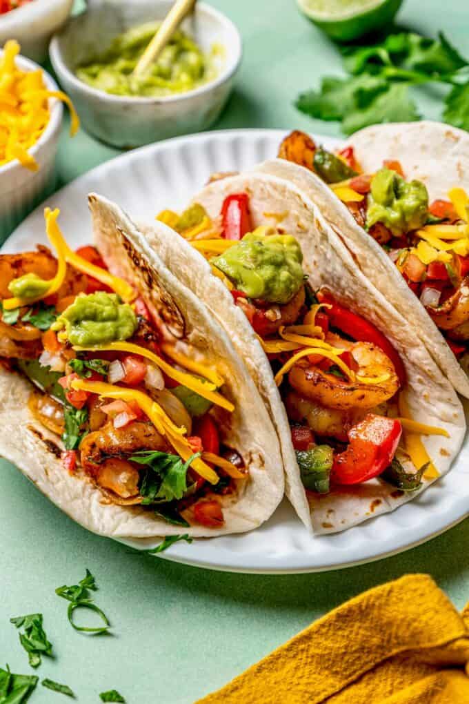 Three shrimp fajitas on a white plate with cheese and avocado on top and extra toppings in the background.