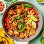 A skillet with cooked, charred shrimp, peppers and onion with cilantro sprinkled on top and lime to the side and surrounded by small bowls of toppings and a text overlay on the photo.