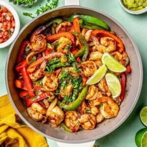 A skillet full of shrimp fajitas with lime wedges and cilantro on top and toppings nearby.