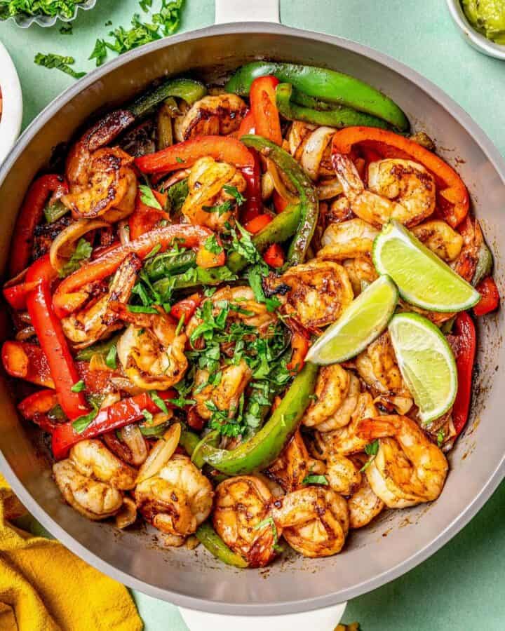 A skillet full of shrimp fajitas with lime wedges and cilantro on top and toppings nearby.