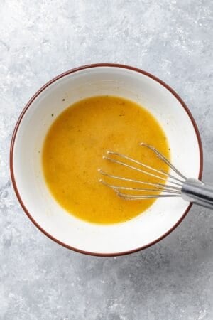 A whisk resting in a small bowl of a salad dressing.