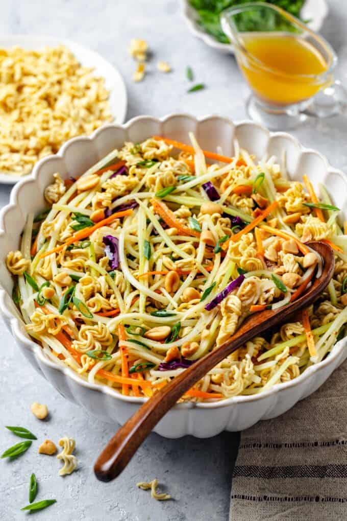 A wooden spoon resting in a large serving bowl full of a ramen salad with broccoli slaw and peanuts.