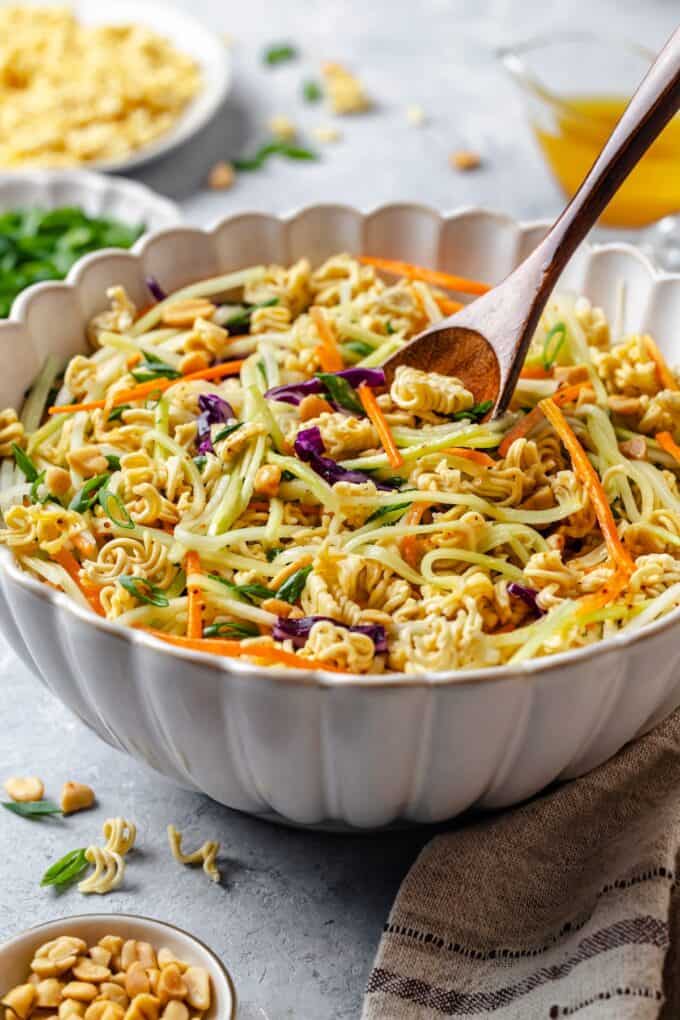 A wooden spoon scooping up a ramen noodle slaw from a large serving bowl.
