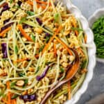 A wooden spoon resting in a large bowl of Asian style coleslaw with peanuts and ramen and a text overlay on the photo.