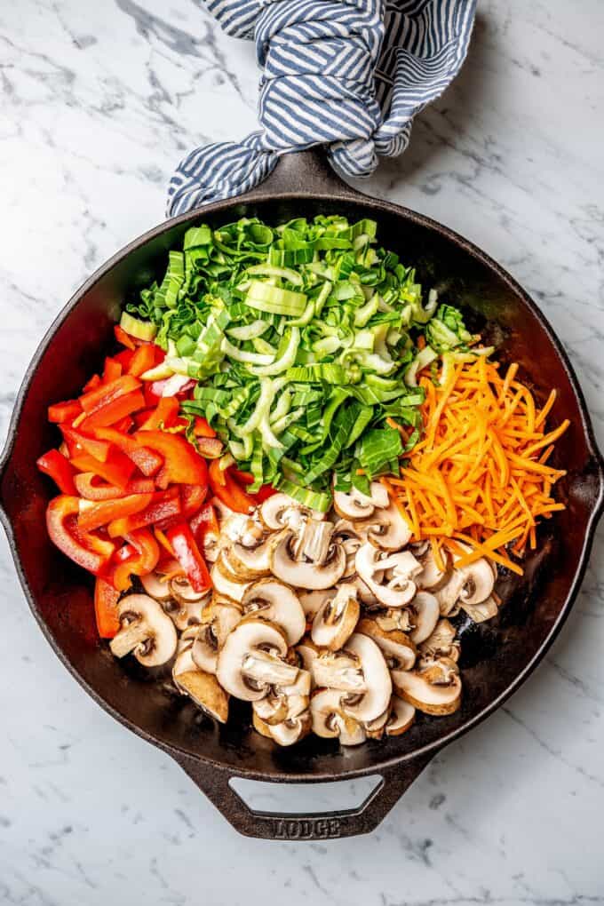 Separate piles of vegetables in a cast iron skillet, including mushrooms, red bell pepper, bok choy and shredded carrots.